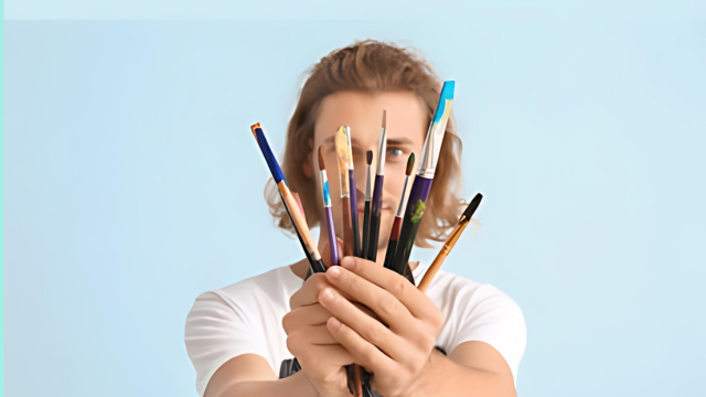 “Artist holding an assortment of drawing pencils and paintbrushes in front of obscured face against a light blue background.”