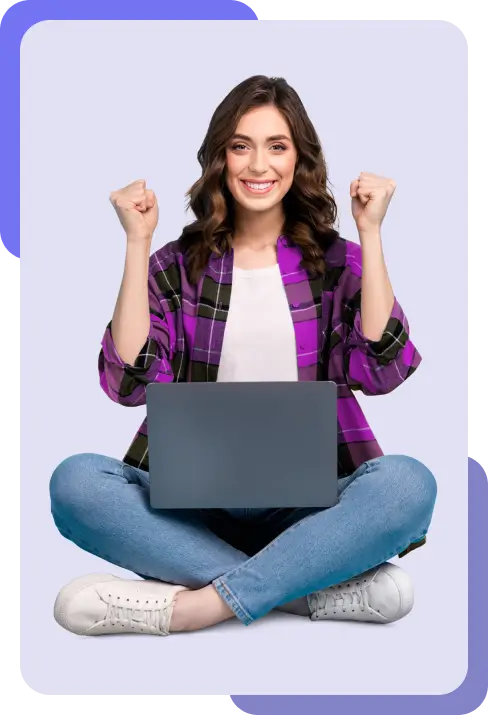 Person celebrating with fists raised while sitting cross-legged with a laptop on their lap, wearing a purple and black checkered shirt, blue jeans, and white sneakers